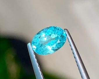 May include: A light blue oval gemstone held in tweezers. The gemstone is transparent and has a slight sparkle.