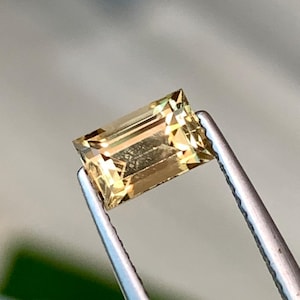 Puede incluir: Una piedra preciosa rectangular facetada de color amarillo dorado. La piedra preciosa se sostiene con unas pinzas.