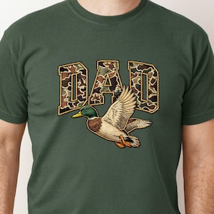 May include: Dark green t-shirt featuring the word "DAD" in a camouflage pattern with a gold outline. A detailed illustration of a flying mallard duck is below the text. The shirt is a short-sleeved crew neck.