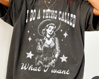 May include: A charcoal gray short-sleeve t-shirt featuring a white graphic print of a cowgirl holding a revolver. The text reads I DO A THING CALLED What I want, surrounded by star icons. The shirt has a crew neckline and a loose, relaxed fit.