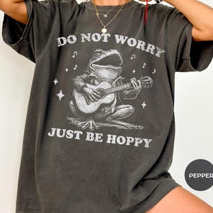 May include: Dark grey t-shirt featuring a frog playing a guitar with the text "DO NOT WORRY JUST BE HOPPY". The graphic is in white and includes musical notes and stars. The shirt is a relaxed fit.