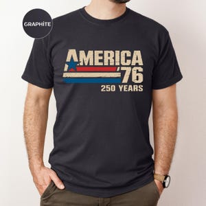 May include: Dark grey t-shirt featuring the text "AMERICA '76 250 YEARS" in a retro design. The graphic includes a blue star, red and blue stripes, and distressed lettering, commemorating the nation's anniversary.