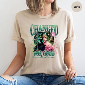 K&ouml;nnte beinhalten: Sandfarbenes T-Shirt mit einem Grafikdesign aus dem Musical "Wicked". Das Design enth&auml;lt den Text "CHANGED FOR GOOD" und "TOGETHER WE'RE UNLIMITED" mit Illustrationen der beiden Hauptfiguren.
