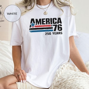 May include: White t-shirt featuring the text "AMERICA '76 250 YEARS" in black, red, blue, and white. The design includes a blue star and horizontal stripes. The shirt is a casual style.