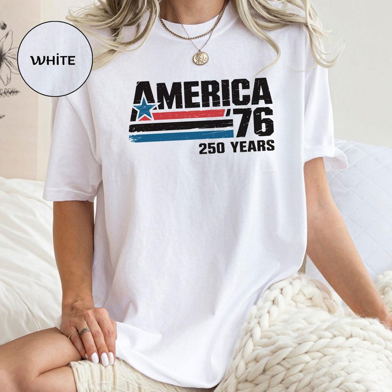 May include: White t-shirt featuring the text "AMERICA '76 250 YEARS" in black, red, blue, and white. The design includes a blue star and horizontal stripes. The shirt is a casual style.