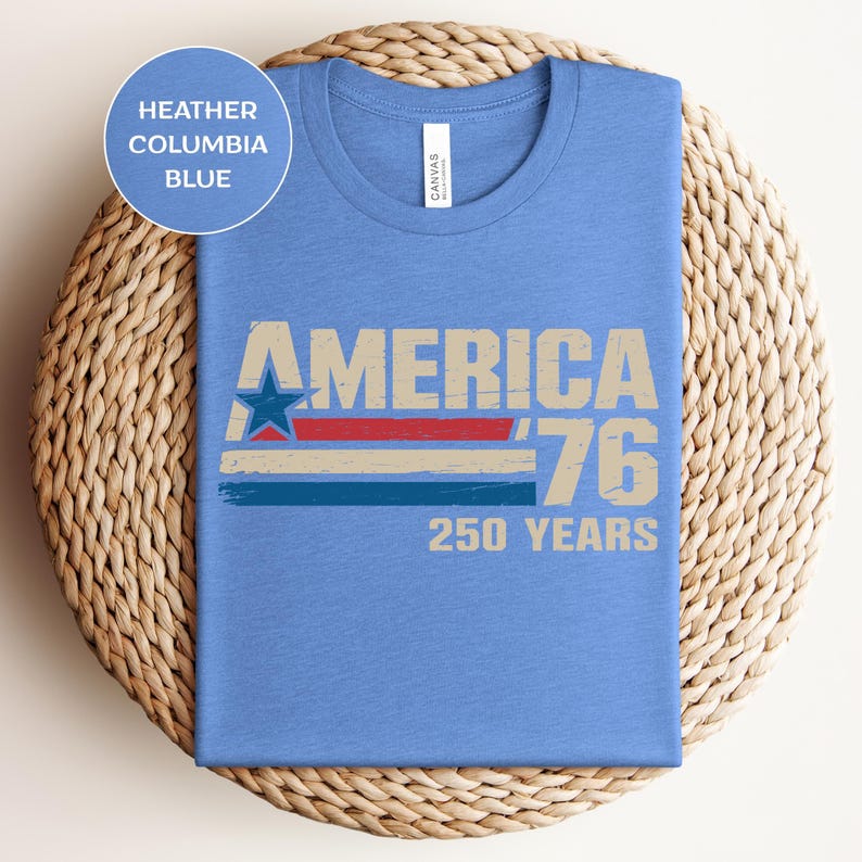 May include: Heather Columbia blue t-shirt featuring the text "AMERICA '76 250 YEARS" in a retro design. The graphic includes a star and stripes in red, white, and blue. The shirt is folded on a woven mat.