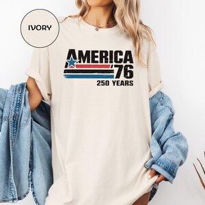 May include: An ivory-coloured t-shirt featuring the text "AMERICA '76 250 YEARS" in black, red, and blue. The design incorporates a star and stripes, commemorating the 250th anniversary. A casual, everyday wear item.