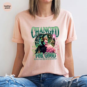 K&ouml;nnte beinhalten: Hellpfirsichfarbenes T-Shirt mit einem grafischen Design. Das Design zeigt den Text "CHANGED FOR GOOD" und "TOGETHER WE'RE UNLIMITED". Die Grafik zeigt zwei Charaktere, einen in einem schwarzen Hexenoutfit und einen in einem rosa Kleid.