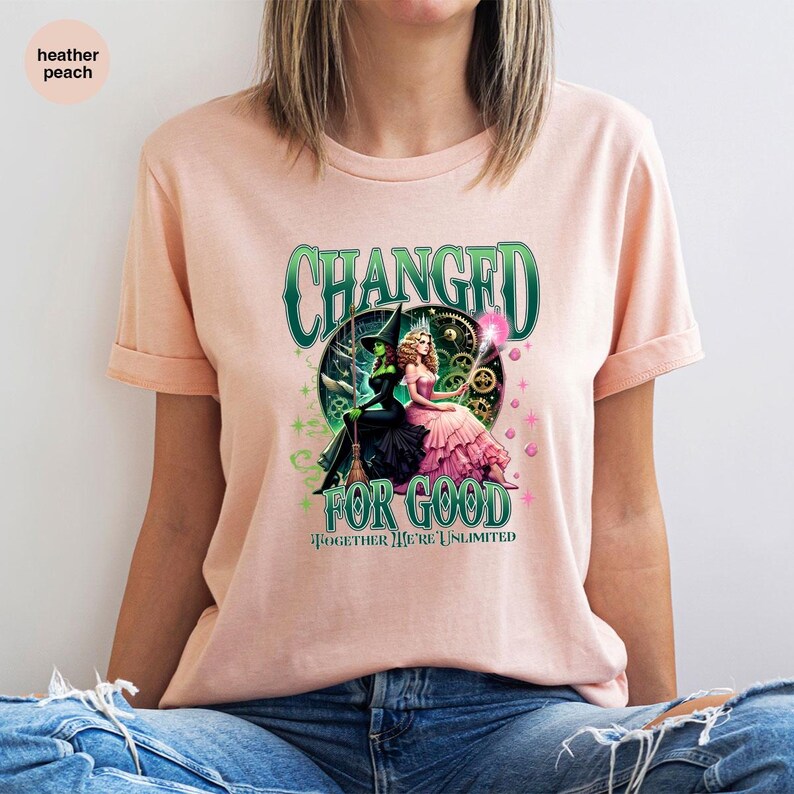K&ouml;nnte beinhalten: Hellpfirsichfarbenes T-Shirt mit einem grafischen Design. Das Design zeigt den Text "CHANGED FOR GOOD" und "TOGETHER WE'RE UNLIMITED". Die Grafik zeigt zwei Charaktere, einen in einem schwarzen Hexenoutfit und einen in einem rosa Kleid.