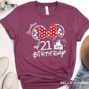 May include: Burgundy t-shirt with a white graphic design. The design includes a red polka dot bow, Mickey Mouse ears, the number 21, a castle, and the words "Jennifer" and "My 21st Birthday". The shirt is from Bella + Canvas.