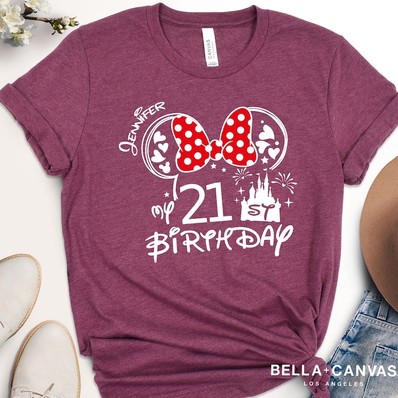 May include: Burgundy t-shirt with a white graphic design. The design includes a red polka dot bow, Mickey Mouse ears, the number 21, a castle, and the words "Jennifer" and "My 21st Birthday". The shirt is from Bella + Canvas.