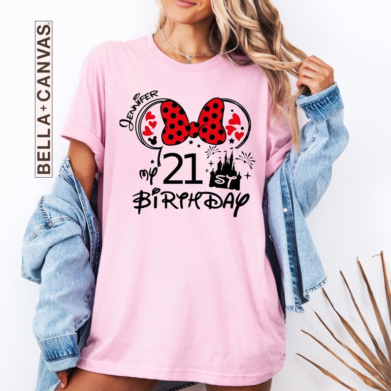 May include: Pink t-shirt with a Minnie Mouse-inspired design. The design includes a red polka dot bow, hearts, the number "21", a castle, and the words "Jennifer" and "Birthday". The shirt is from Bella+Canvas.