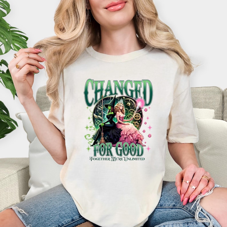 K&ouml;nnte beinhalten: Cremefarbenes T-Shirt mit einer Grafik von zwei Frauen in Fantasy-Kleidung, eine in Gr&uuml;n und Schwarz, die andere in Rosa. Der Text "CHANGED FOR GOOD" und "TOGETHER WE'RE UNLIMITED" ist in Gr&uuml;n und Schwarz dargestellt.