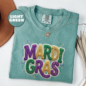 May include: Light green Comfort Colors t-shirt with the words "Mardi Gras" embroidered in purple, gold, and green. The shirt has a crew neck and short sleeves. A silver necklace with a pendant is draped on the shirt.