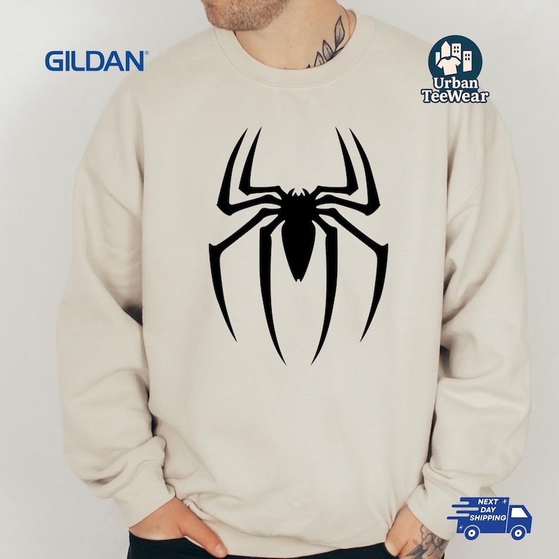 May include: A cream-coloured sweatshirt with a large black spider design on the front. The sweatshirt has long sleeves and a crew neck. The brand name "GILDAN" is visible on the upper left. The words "Urban TeeWear" are on the upper right.