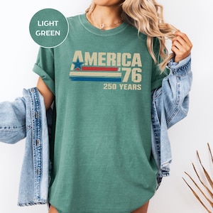 May include: Light green t-shirt featuring the text "AMERICA '76 250 YEARS". The design includes a blue star, red and blue stripes, and distressed details, celebrating the 250th anniversary of America.