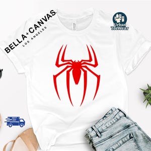 May include: White t-shirt with a large red spider graphic. The sleeve has the text "BELLA + CANVAS LOS ANGELES" and "Urban TeeWear" is printed on the upper right. The shirt is paired with blue jeans.