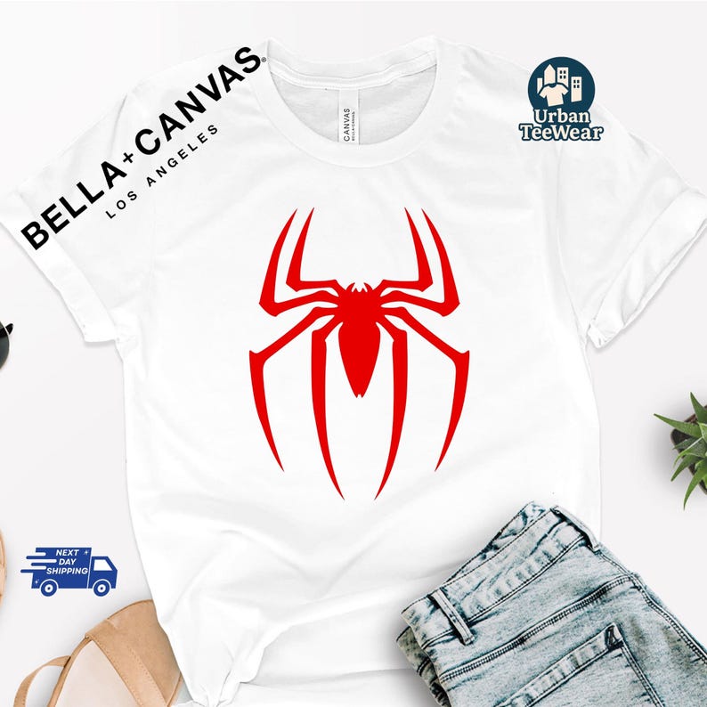 May include: White t-shirt with a large red spider graphic. The sleeve has the text "BELLA + CANVAS LOS ANGELES" and "Urban TeeWear" is printed on the upper right. The shirt is paired with blue jeans.