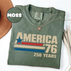 May include: A moss green t-shirt with the text "AMERICA '76 250 YEARS" in a distressed font. The design includes a blue star, red stripe, and blue stripe. A silver necklace with a pendant is draped on the shirt.