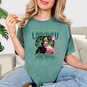 K&ouml;nnte beinhalten: Ein teal-gr&uuml;nes T-Shirt mit einer Grafik von zwei Frauen, eine in einem schwarzen Hexenkost&uuml;m und die andere in einem rosa Kleid. Der Text "CHANGED FOR GOOD" und "TOGETHER WE'RE UNLIMITED" ist auf dem Shirt aufgedruckt.