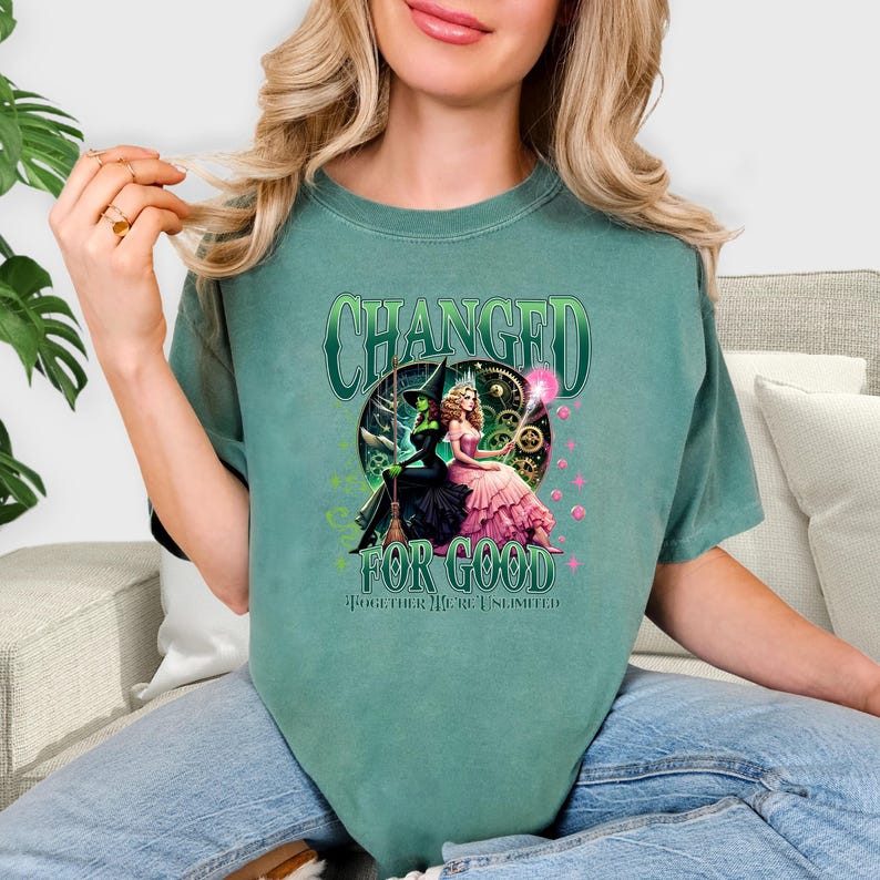 K&ouml;nnte beinhalten: Ein teal-gr&uuml;nes T-Shirt mit einer Grafik von zwei Frauen, eine in einem schwarzen Hexenkost&uuml;m und die andere in einem rosa Kleid. Der Text "CHANGED FOR GOOD" und "TOGETHER WE'RE UNLIMITED" ist auf dem Shirt aufgedruckt.