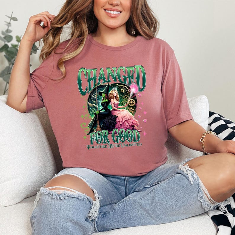 K&ouml;nnte beinhalten: Mauvefarbenes T-Shirt mit einem Grafikdesign aus dem Musical "Wicked". Das Design zeigt die Worte "CHANGED" und "FOR GOOD" in Gr&uuml;n, zusammen mit Bildern der beiden Hauptfiguren. Der Satz "TOGETHER WE'RE UNLIMITED" ist ebenfalls enthalten.