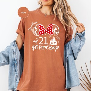 May include: A terracotta-colored t-shirt with a white graphic featuring a red polka-dot bow, hearts, and a castle. The text reads "Jennifer", "my 21st Birthday". The shirt has rolled-up sleeves and a relaxed fit.