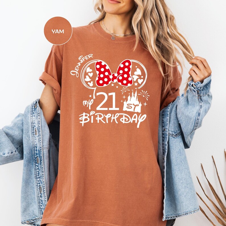 May include: A terracotta-colored t-shirt with a white graphic featuring a red polka-dot bow, hearts, and a castle. The text reads "Jennifer", "my 21st Birthday". The shirt has rolled-up sleeves and a relaxed fit.