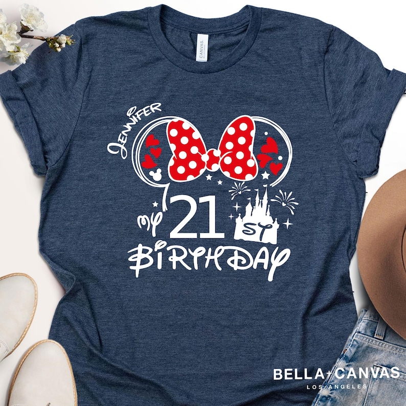 May include: Navy blue t-shirt with a white graphic design. The design includes a red polka dot bow, Mickey Mouse ears, hearts, a castle, fireworks, and the text "Jennifer", "21", and "Birthday". The shirt is from Bella + Canvas, Los Angeles.