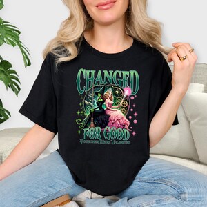 K&ouml;nnte beinhalten: Schwarzes T-Shirt mit einem grafischen Design. Das Design enth&auml;lt den Text "CHANGED FOR GOOD" in Gr&uuml;n und eine Illustration von zwei Figuren. Eine ist eine gr&uuml;n geh&auml;utete Hexe, die andere eine Frau in einem rosa Kleid. Der Text "TOGETHER WE'RE UNLIMITED" ist ebenfalls enthalten.