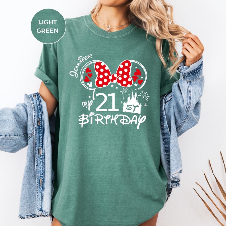 May include: Light green t-shirt with a graphic design for a 21st birthday. The design includes a red polka dot bow, hearts, a castle, and the text "Jennifer" and "my 21st Birthday" in white. The shirt is a solid color.