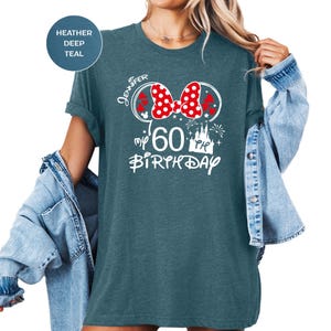 Puede incluir: Una camiseta de color verde azulado con un dise&ntilde;o gr&aacute;fico blanco que presenta un lazo rojo de lunares, el nombre "Jennifer", el n&uacute;mero "60" y las palabras "my birthday". La camiseta tiene las mangas enrolladas.