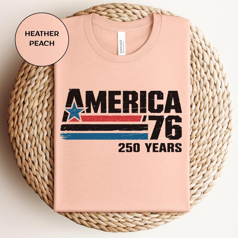 May include: Peach-coloured t-shirt with the text "AMERICA '76 250 YEARS" in black. The design includes a blue star and red and blue stripes. The shirt is folded on a woven, beige surface.