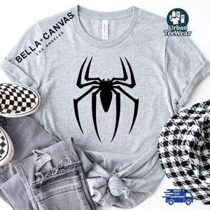 May include: A light grey t-shirt with a large black spider design. The shirt has the text "BELLA + CANVAS LOS ANGELES" printed on the upper left side. The shirt is paired with grey jeans and checkered shoes.