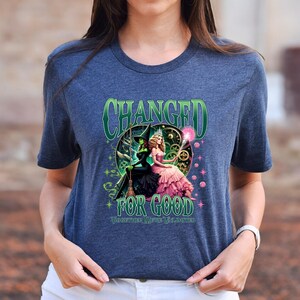 K&ouml;nnte beinhalten: Ein marineblaues T-Shirt mit einer Grafik aus dem Musical "Wicked". Das Design enth&auml;lt den Text "CHANGED FOR GOOD" in Gr&uuml;n, mit Bildern von Elphaba und Glinda. Das T-Shirt hat einen Rundhalsausschnitt und kurze &Auml;rmel.