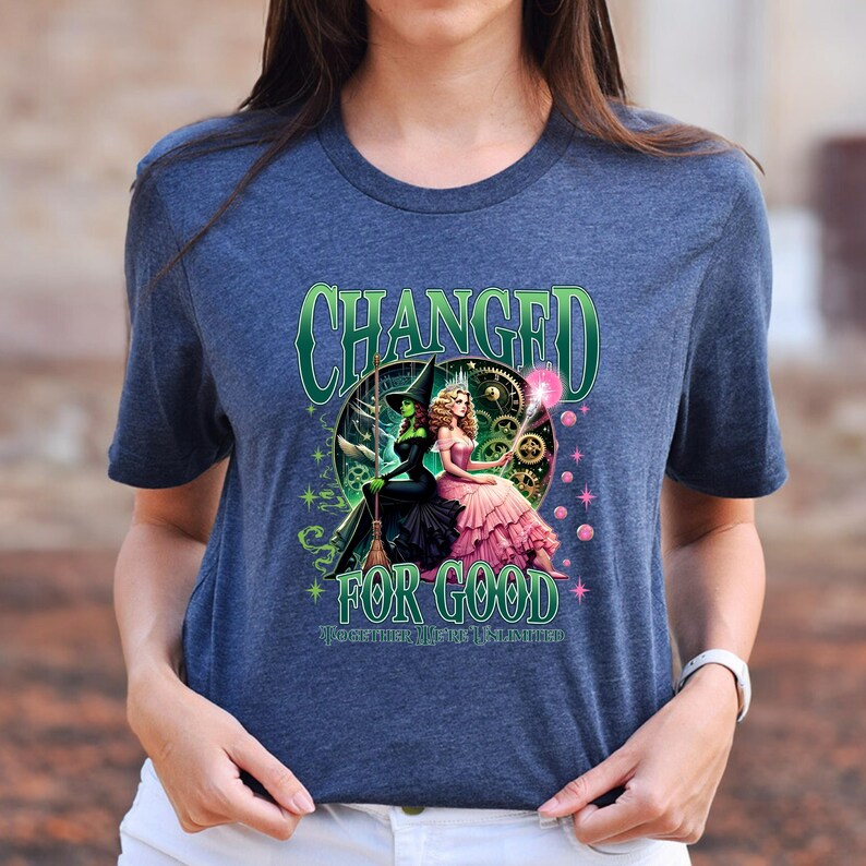 K&ouml;nnte beinhalten: Ein marineblaues T-Shirt mit einer Grafik aus dem Musical "Wicked". Das Design enth&auml;lt den Text "CHANGED FOR GOOD" in Gr&uuml;n, mit Bildern von Elphaba und Glinda. Das T-Shirt hat einen Rundhalsausschnitt und kurze &Auml;rmel.