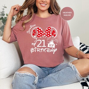 May include: Heather mauve t-shirt with a white graphic. The graphic includes a red polka dot bow, hearts, the number "21", a castle, fireworks, and the word "Birthday". The name "Jennifer" is also included in the graphic.