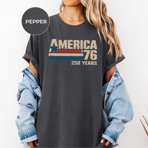 May include: Dark grey t-shirt featuring the text "AMERICA '76 250 YEARS" in a retro design. The graphic includes a blue star, red and white stripes, and the number 76. The shirt is worn with a denim jacket.