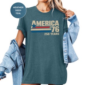 May include: Deep teal t-shirt featuring the text "AMERICA '76 250 YEARS" in a vintage style. The design includes a blue star, red and blue stripes, and a beige background. The shirt has a relaxed fit.