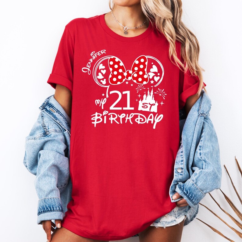 May include: Red t-shirt with a white graphic featuring a polka-dot bow, hearts, the number "21", a castle, and the words "Jennifer" and "Birthday". The shirt has short sleeves.