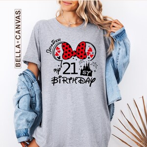 May include: A gray t-shirt with a Minnie Mouse-themed design for a 21st birthday. The design includes a red polka-dot bow, hearts, a castle, fireworks, and the name "Jennifer". The shirt is from Bella+Canvas.