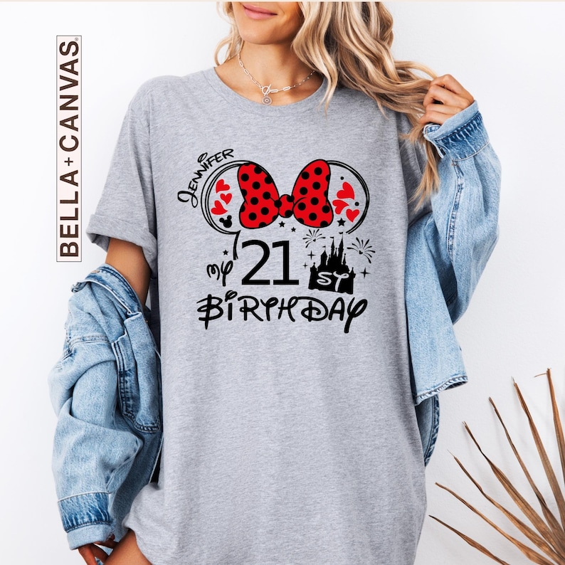 May include: A gray t-shirt with a Minnie Mouse-themed design for a 21st birthday. The design includes a red polka-dot bow, hearts, a castle, fireworks, and the name "Jennifer". The shirt is from Bella+Canvas.