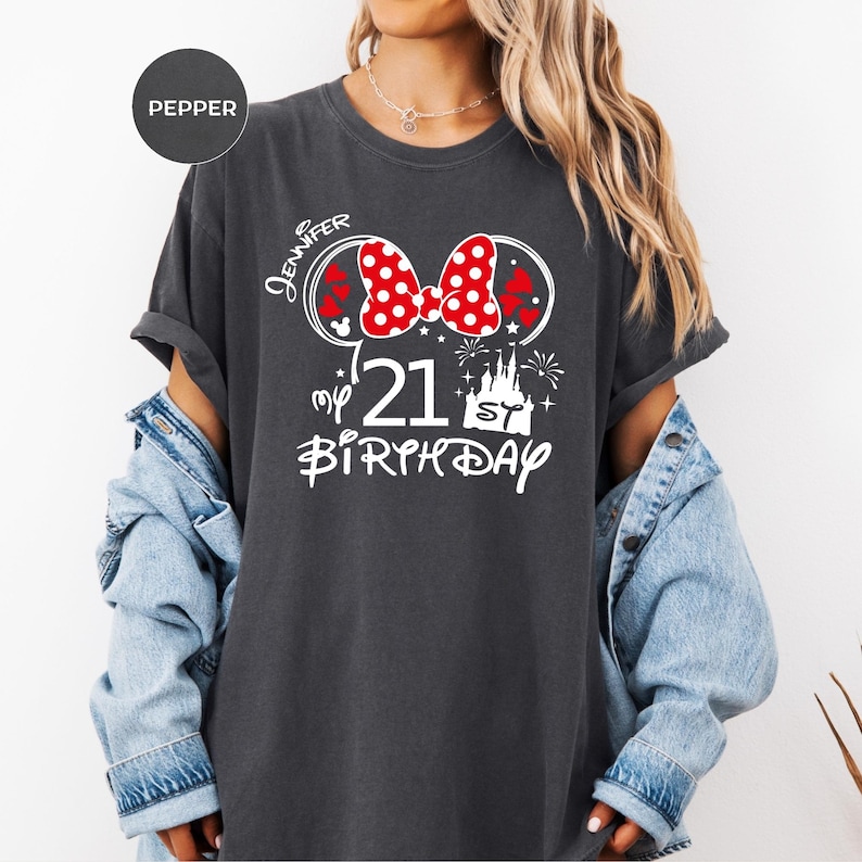May include: Dark gray t-shirt with a Minnie Mouse-themed design. The design includes a red polka dot bow, the name "Jennifer", the number "21", the words "my birthday", and a castle graphic.