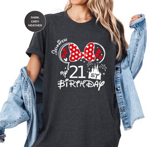 May include: Dark grey heather t-shirt with a Minnie Mouse-themed design for a 21st birthday. The design includes a red bow with white polka dots, hearts, and the name "Jennifer". The text "my 21st Birthday" is also featured.