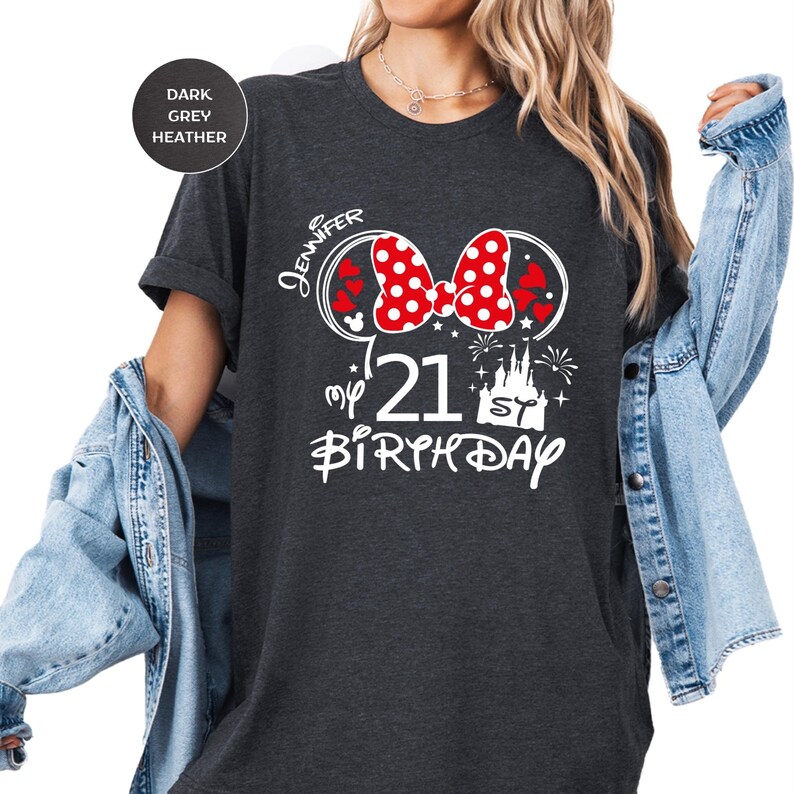 May include: Dark grey heather t-shirt with a Minnie Mouse-themed design for a 21st birthday. The design includes a red bow with white polka dots, hearts, and the name "Jennifer". The text "my 21st Birthday" is also featured.
