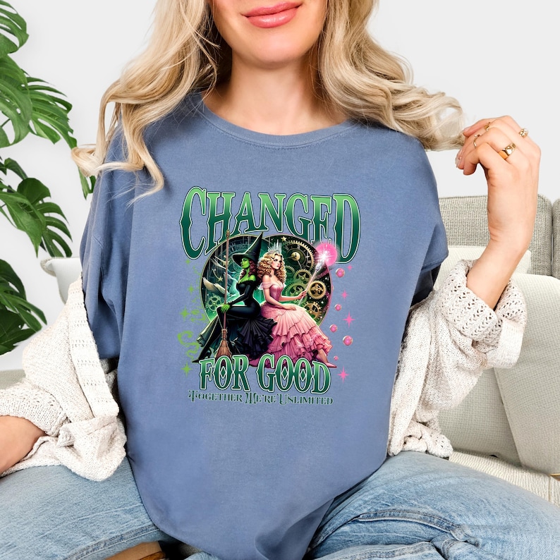 K&ouml;nnte beinhalten: Blaues T-Shirt mit einem Grafikmotiv aus dem Musical "Wicked". Das Design zeigt den Text "CHANGED FOR GOOD" in Gr&uuml;n sowie Illustrationen der beiden Hauptfiguren, Elphaba und Glinda.