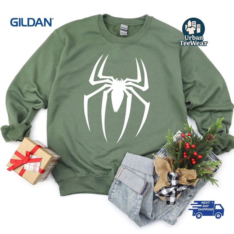 May include: A sage green sweatshirt with a large white spider graphic. The sweatshirt is from the brand Gildan and features the Urban TeeWear logo. The image includes a wrapped gift, jeans, and holiday greenery.