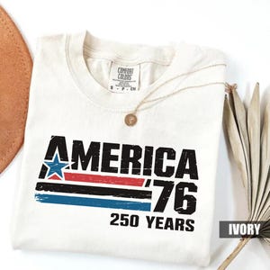 May include: Cream-coloured t-shirt featuring the text "AMERICA '76 250 YEARS" in black, red, blue, and white. The design includes a star and stripes. The shirt has a round neckline and a gold necklace draped across it.