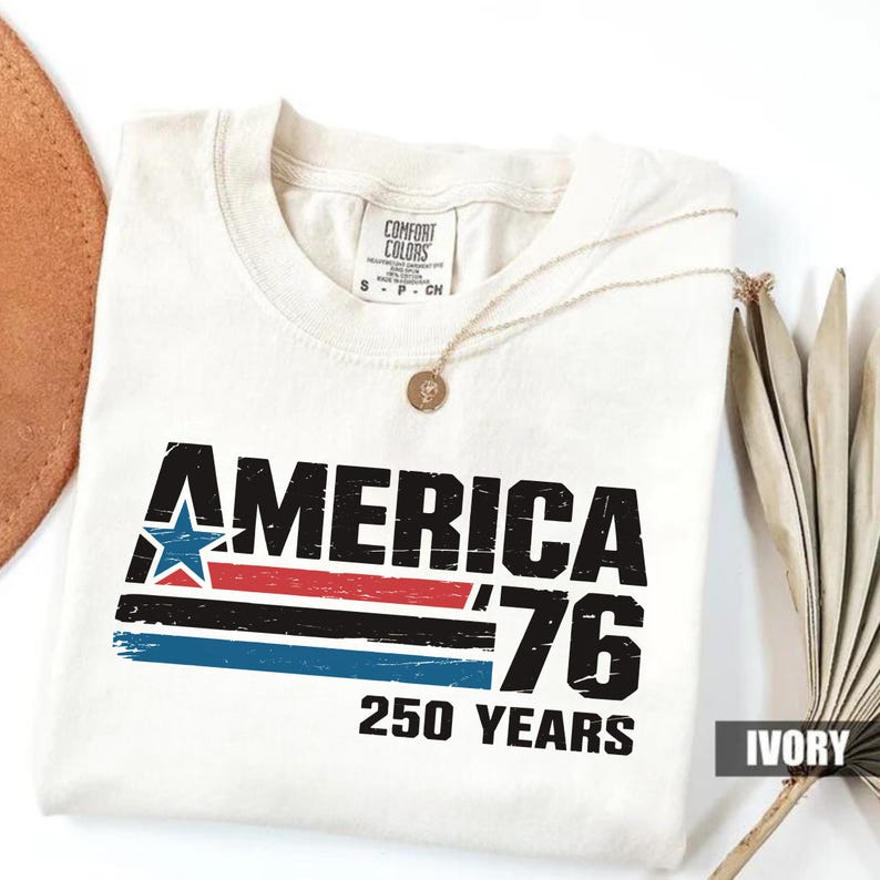 May include: Cream-coloured t-shirt featuring the text "AMERICA '76 250 YEARS" in black, red, blue, and white. The design includes a star and stripes. The shirt has a round neckline and a gold necklace draped across it.