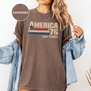 May include: Espresso-coloured t-shirt featuring the text "AMERICA '76 250 YEARS" in a vintage style. The design includes a blue star and red and blue stripes. The shirt has rolled-up sleeves and is paired with a denim jacket.